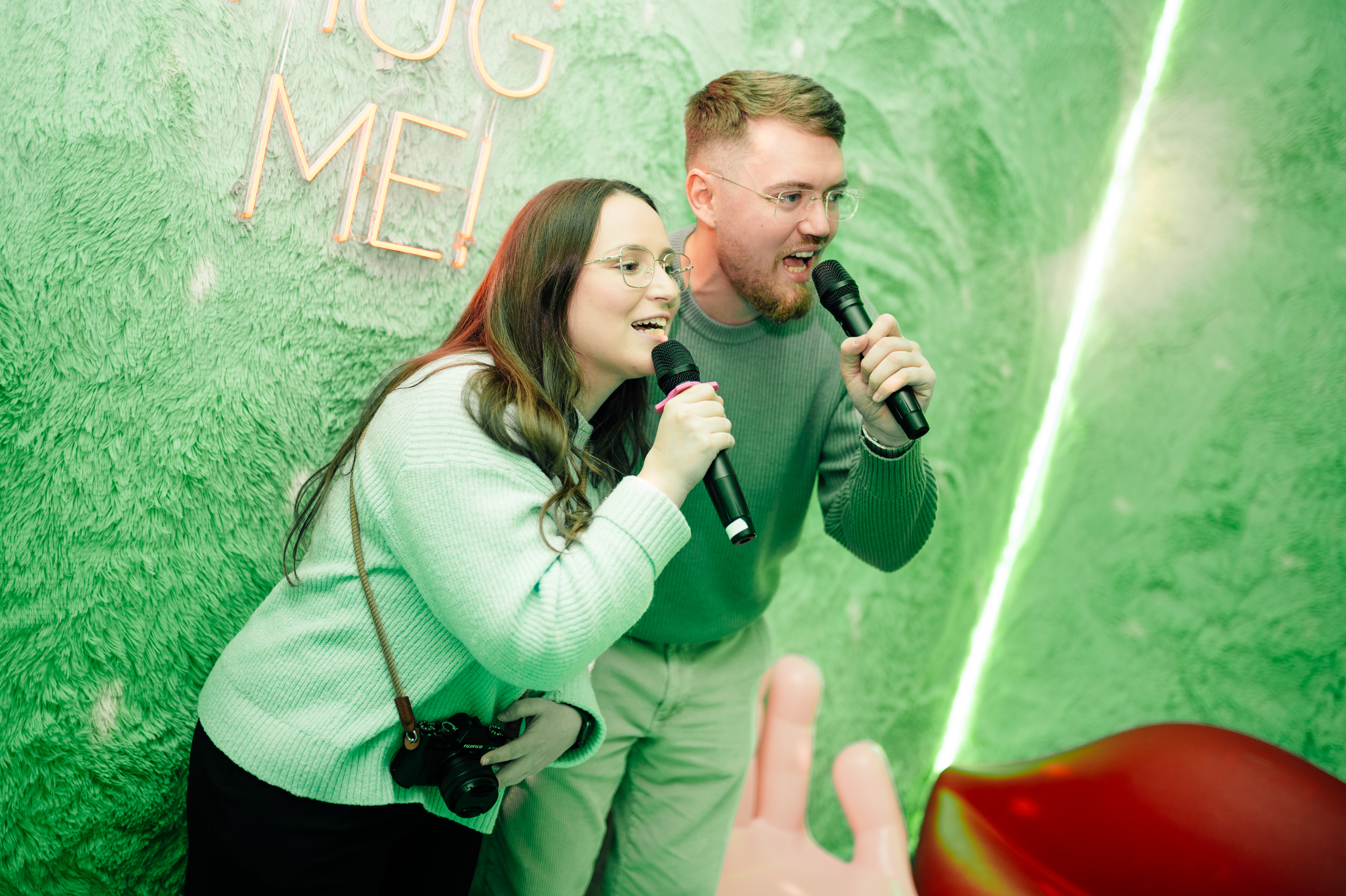 Zwei junge Erwachsene singen in Mikrofone in einer Kabine mit flauschigen Wänden